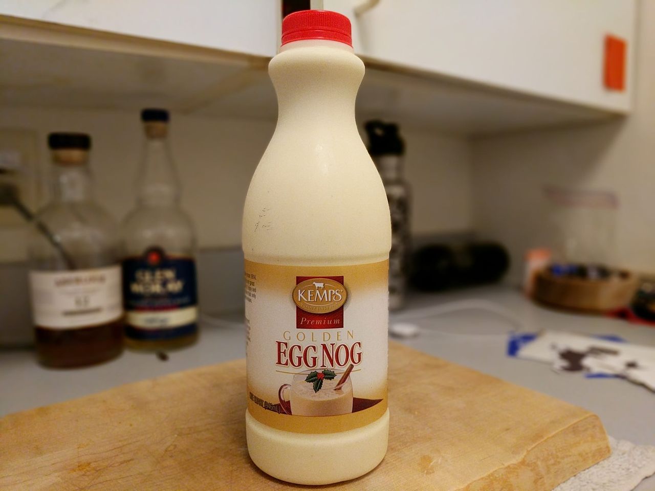 A bottle of Kemps Premium Golden Egg Nog on a cutting board in front of liquor bottles
