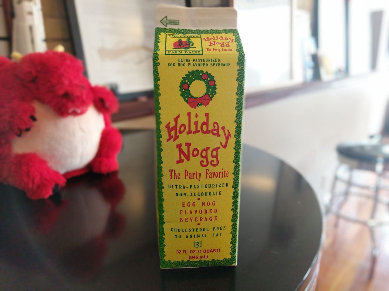 A carton of Twin Pines Holiday Nogg, “The Party Favorite”, next to a red plush toy